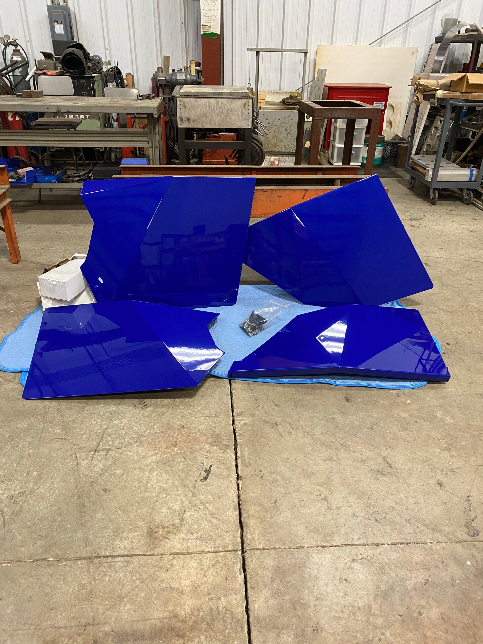Four large, shiny, deep blue Rzr XP 4 1000 doors with angular shapes rest on a blue blanket on a workshop floor surrounded by industrial machines, workbenches, and tools in a spacious workshop with concrete flooring and white walls.
