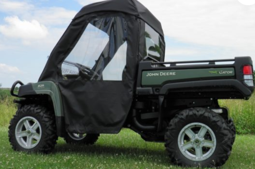 A John Deere Gator 550/560/590 with a Full Cab for Hard Windshield, featuring large off-road tires and an enclosed black soft cover with clear windows, is parked on grass under a partly cloudy sky, viewed from the side.