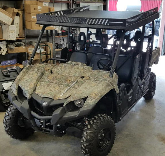 A Yamaha Viking 6 (Vi) with a metal roof, camo paint, large off-road tires, and seating for four is parked indoors on a concrete floor, surrounded by shelves with boxes and equipment in the background.