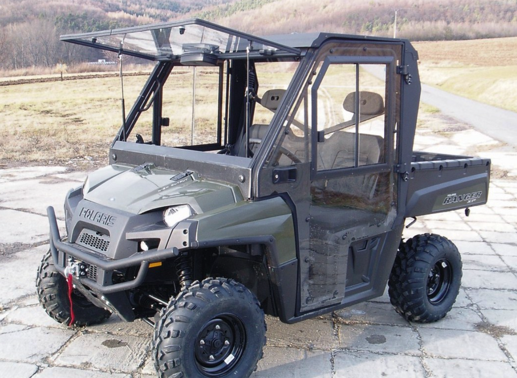 A Polaris Ranger XP800 / Ranger 570 FULL SIZE Cab Kit-equipped UTV with a hard roof and transparent doors is parked on cracked pavement, featuring rugged tires and a covered cab for off-road use. Fields, trees, and hills appear in the background.
