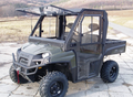 A Polaris Ranger XP800 / Ranger 570 FULL SIZE Cab Kit-equipped UTV with a hard roof and transparent doors is parked on cracked pavement, featuring rugged tires and a covered cab for off-road use. Fields, trees, and hills appear in the background.