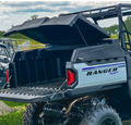 A black and gray Polaris Ranger 570SP fitted with the HIGHLANDS Bed Cover is parked outdoors, its rear cargo bed open to reveal spacious storage. Grass, fencing, and another ATV are visible in the blurred background.