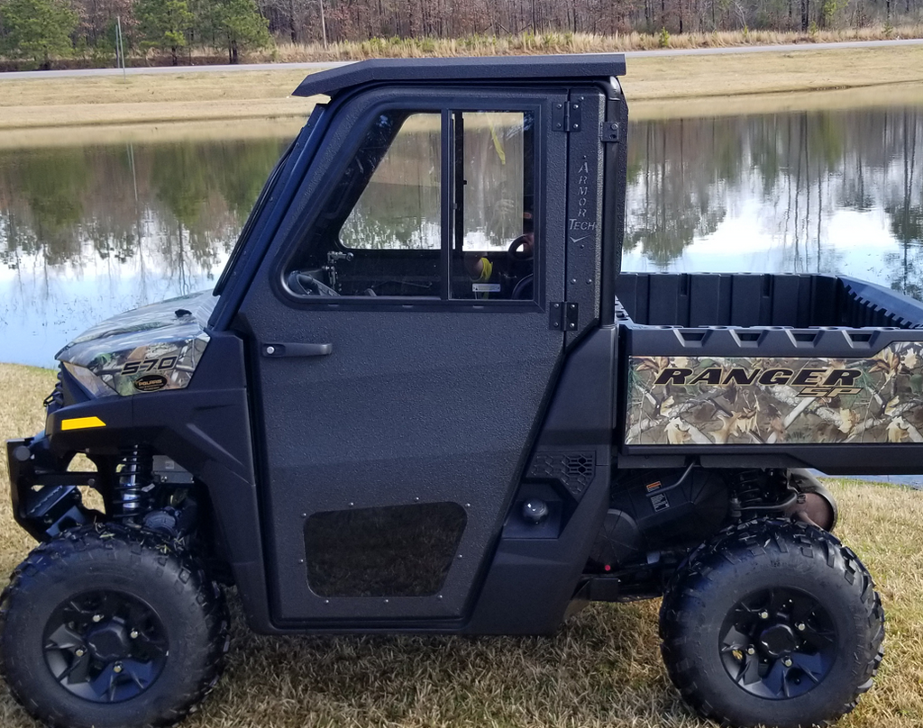 A Polaris Ranger 570 SP with Armor Tech full steel doors and camo rear detailing is parked on grass by a pond, its off-road tires ready for action. A reflection of trees and sky appears in the water behind the black side-by-side utility vehicle.
