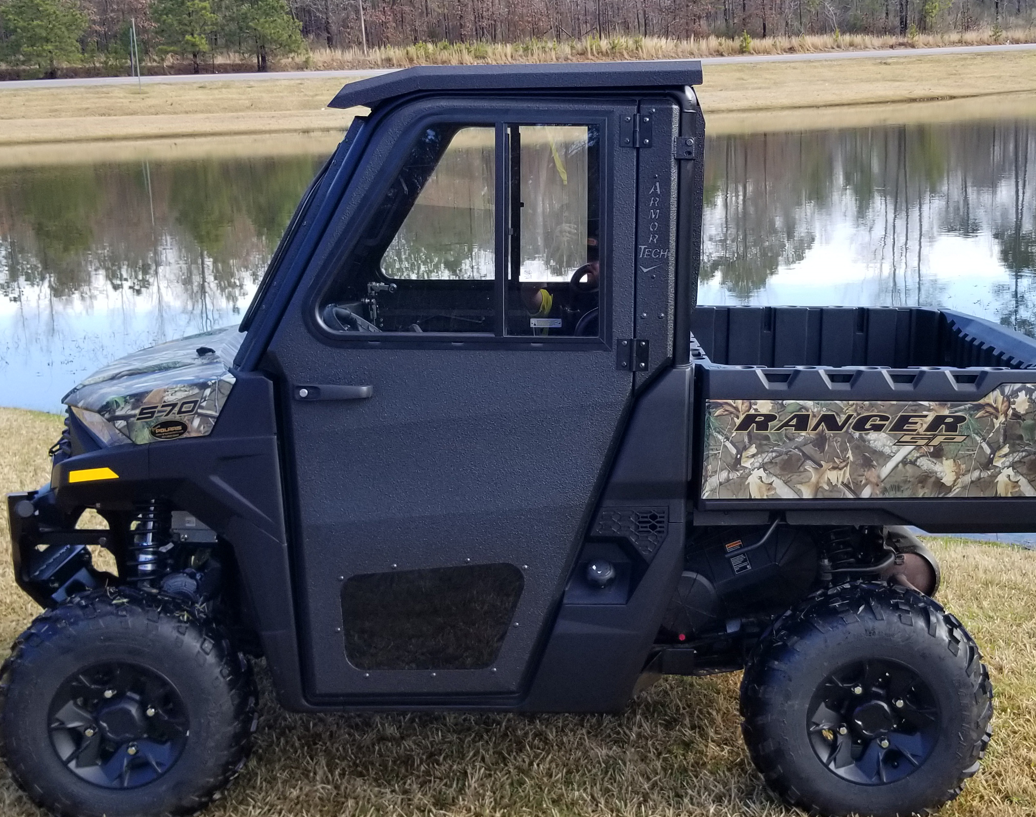 A Polaris Ranger 570 SP with Armor Tech full steel doors and camo rear detailing is parked on grass by a pond, its off-road tires ready for action. A reflection of trees and sky appears in the water behind the black side-by-side utility vehicle.