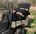 The Yamaha Viking High Seat, featuring a camouflage pattern, is mounted on the cargo bed with a black metal and cushioned bench, armrests, a backrest, and two ladder steps for access. The background shows trees and rocky terrain.