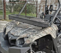 A Yamaha ATV with a camouflage pattern is parked by a fence and metal building. It features a large, rectangular black Yamaha Viking Roll Bar Basket (Bolts On Factory Frame) mounted on the front for carrying cargo.
