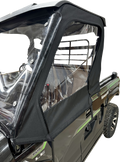 Close-up of a black Kawasaki utility vehicle with a clear plastic and black fabric cab enclosure. The enclosure covers the side window and door area, secured with zippers and fasteners. The rear cargo area and metal rack are visible.