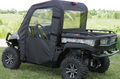 A John Deere XUV 550/560/590 with a Soft Door Rear Window Combo is parked on grass. The black canvas cab enclosure has clear plastic windows, and the cargo bed is visible. Green trees and rolling fields are in the background.