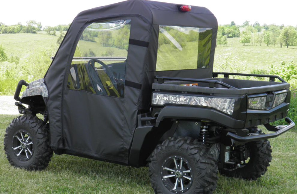 A John Deere XUV 550/560/590 with a Soft Door Rear Window Combo is parked on grass. The black canvas cab enclosure has clear plastic windows, and the cargo bed is visible. Green trees and rolling fields are in the background.