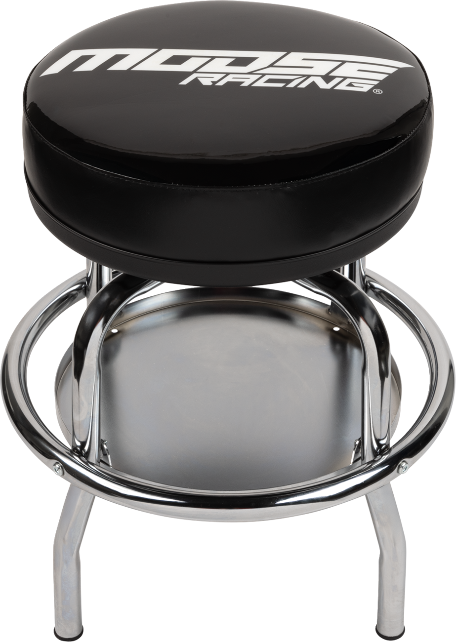 A round black stool with a cushioned seat featuring the white Moose Racing logo. The stool has a shiny chrome frame with four legs, a circular footrest, and a round lower shelf. The design is sturdy and industrial.