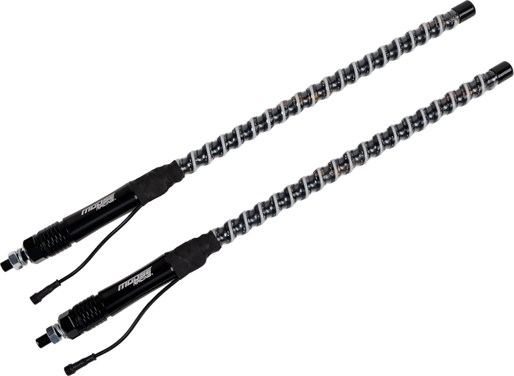 Two black automotive shock absorbers with coiled springs are shown diagonally on a light gray background. The shocks feature connectors and cables at the base, and the springs are metallic with visible orange details along their coils.