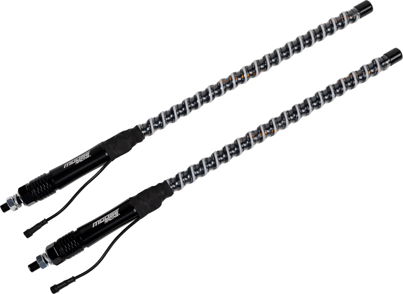 Two black automotive shock absorbers with coiled springs are shown diagonally on a light gray background. The shocks feature connectors and cables at the base, and the springs are metallic with visible orange details along their coils.