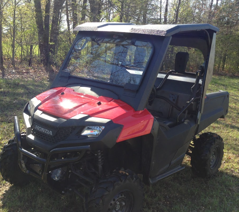 The Honda Pioneer 700, equipped with a front glass windshield, is parked on grass among trees. The red and black side-by-side has a roof, large off-road tires, and an open two-seat cabin as sunlight filters through the scene.