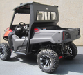 A Honda Pioneer 700 with a rear glass windshield is shown from the side and rear, featuring black and red trim, off-road tires, black wheels, and a partial cab frame. The UTV is parked on pavement next to a beige concrete wall.