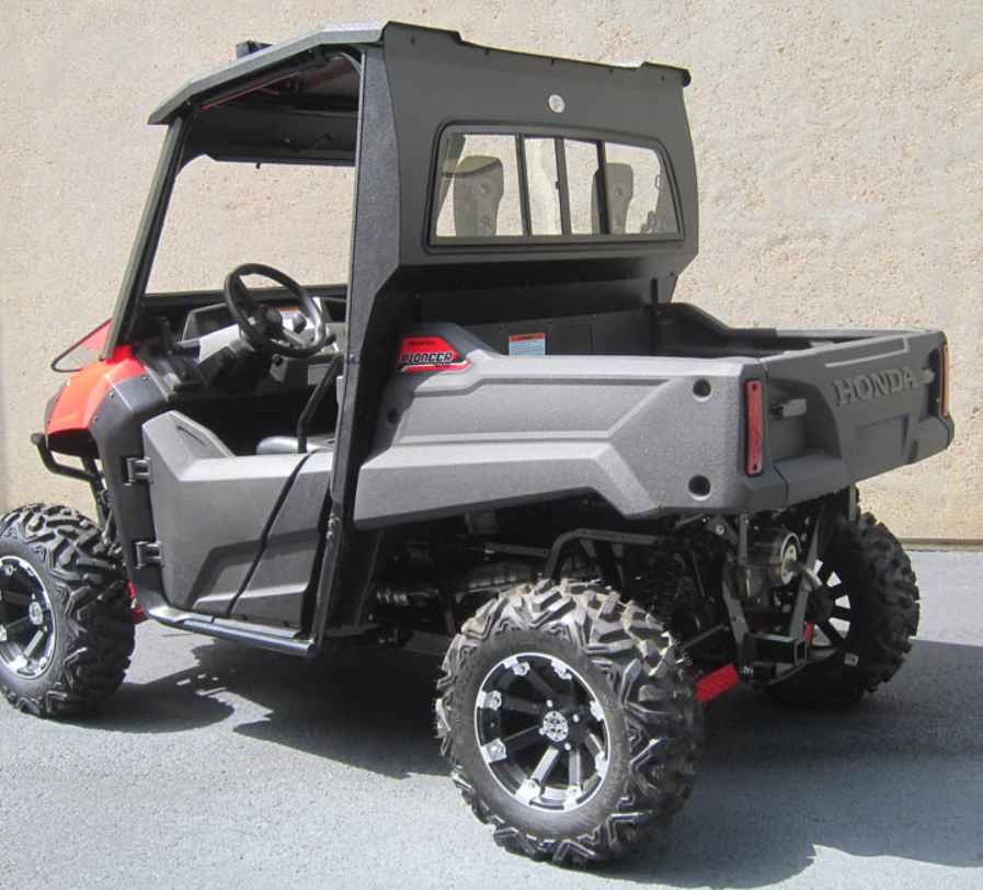 A Honda Pioneer 700 with a rear glass windshield is shown from the side and rear, featuring black and red trim, off-road tires, black wheels, and a partial cab frame. The UTV is parked on pavement next to a beige concrete wall.
