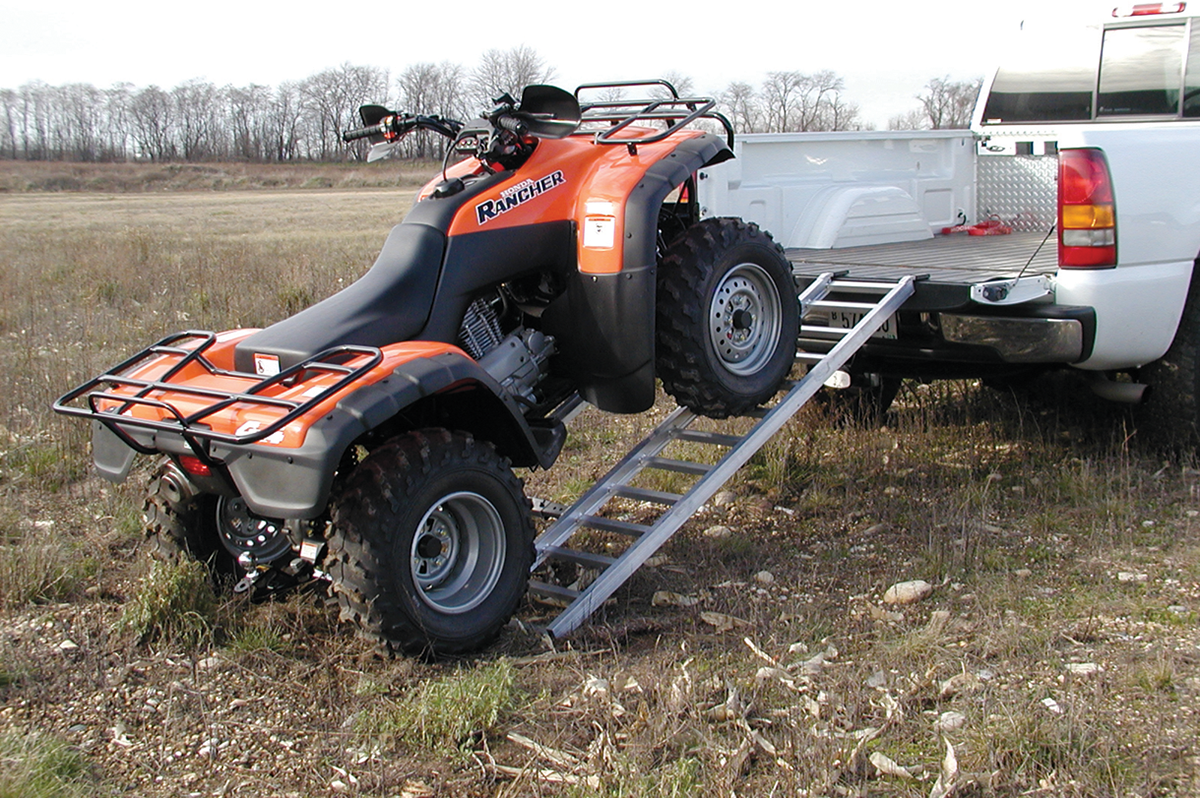 An orange ATV is being loaded into the bed of a white pickup truck using two metal ramps on a grassy field. The ATV is halfway up the ramps, with its front wheels on the truck bed and rear wheels on the ramps. Trees are visible in the background.