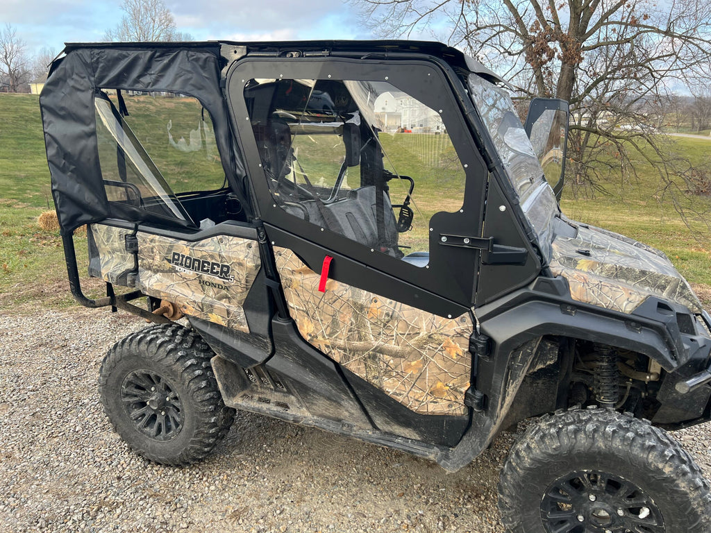 A Honda Pioneer 1000-5 with a camouflage pattern features an enclosure kit: aluminum upper front doors, soft rear doors, plus middle and back soft panels. Its parked on gravel near grass, leafless trees, and a distant white house.