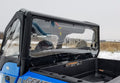 A close-up of a Polaris General Rear Windshield W/Vent-Gp 2016+ installed on a blue side-by-side utility vehicle. The clear panel shows the empty bed and interior cab, with snowy terrain, dry grass, and an overcast sky in the background.
