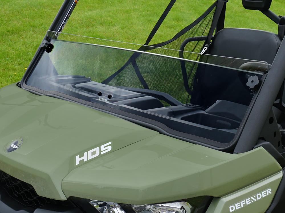 A close-up of a green off-road utility vehicle featuring a Can Am Defender Half Windshield Clear Hard Coated Scratch Resistant 2016+, with mesh netting on the side and two black seats, parked on grass.