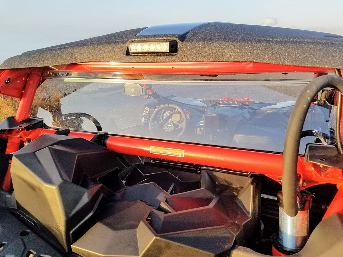 Rear view of a Can Am Maverick X-3 with vented rear windshield (2017+), showing black and red interior, two bucket seats, safety harnesses, roll cage, and steering wheel with dashboard controls illuminated by sunlight.