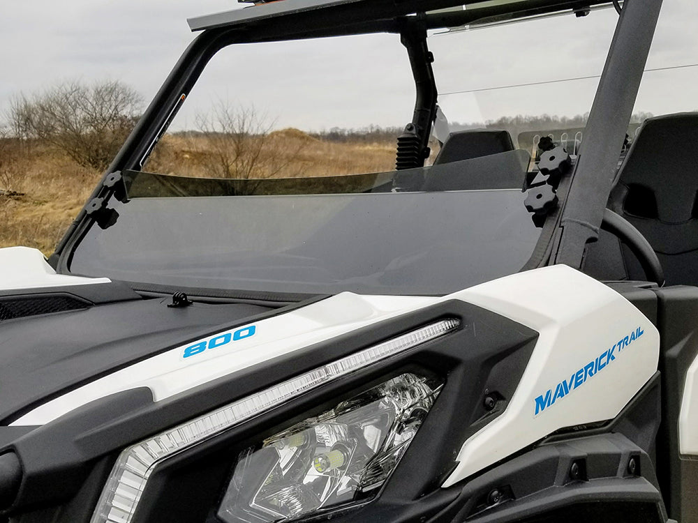 Close-up of a Can Am Maverick Trail with tinted short windshield (2018+), highlighting the windshield and headlight. Maverick 800 is visible on the body, with dry grass and bushes in the overcast background.