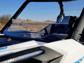 Close-up of a Can Am Maverick Trail Short Windshield (Hard Coated, Scratch Resistant, 2018+) on a white off-road vehicle with black seats and an LED light bar. Dry grass and shrubs appear in the background under a blue sky.