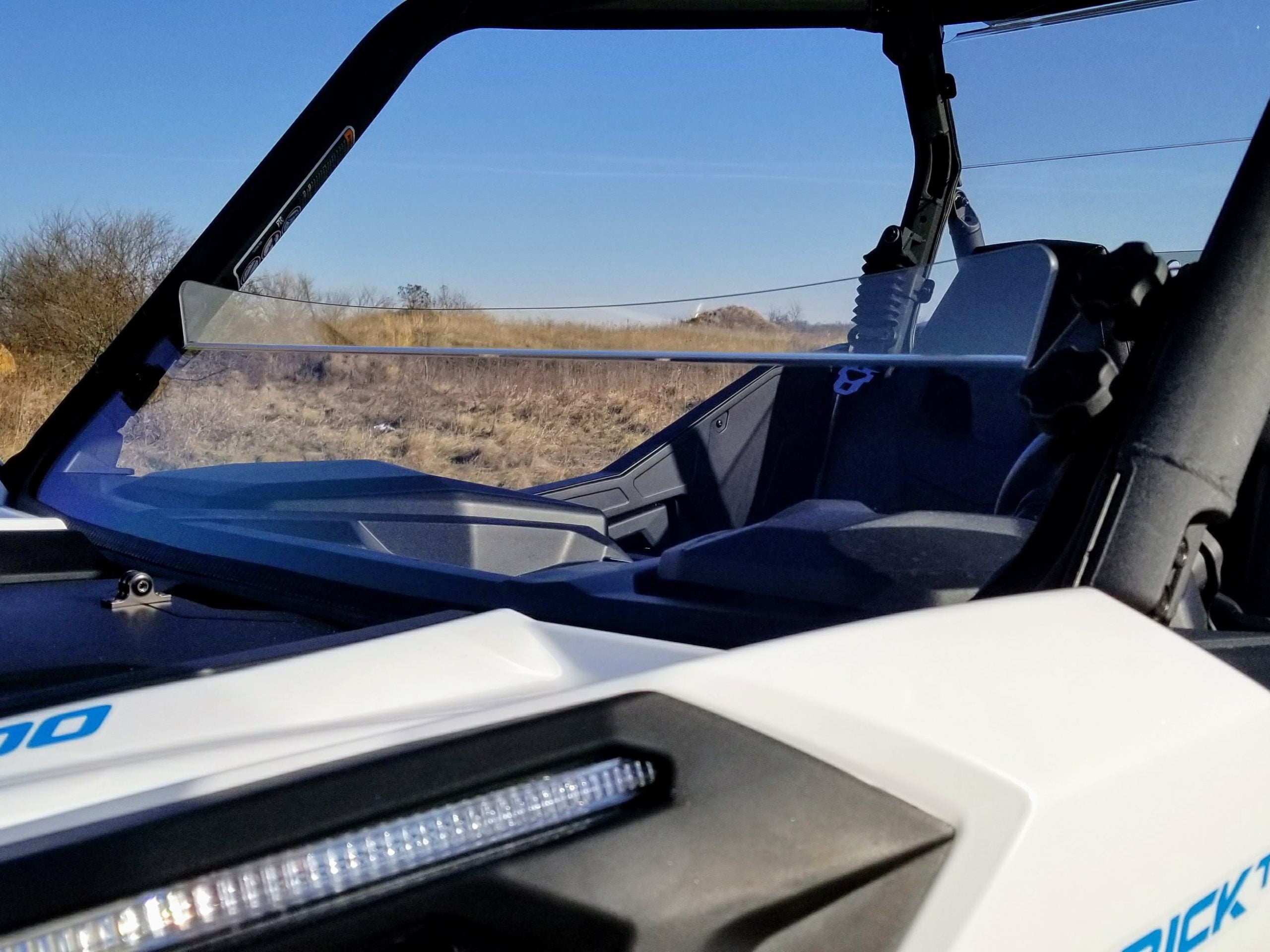 Close-up of a Can Am Maverick Trail Short Windshield (Hard Coated, Scratch Resistant, 2018+) on a white off-road vehicle with black seats and an LED light bar. Dry grass and shrubs appear in the background under a blue sky.