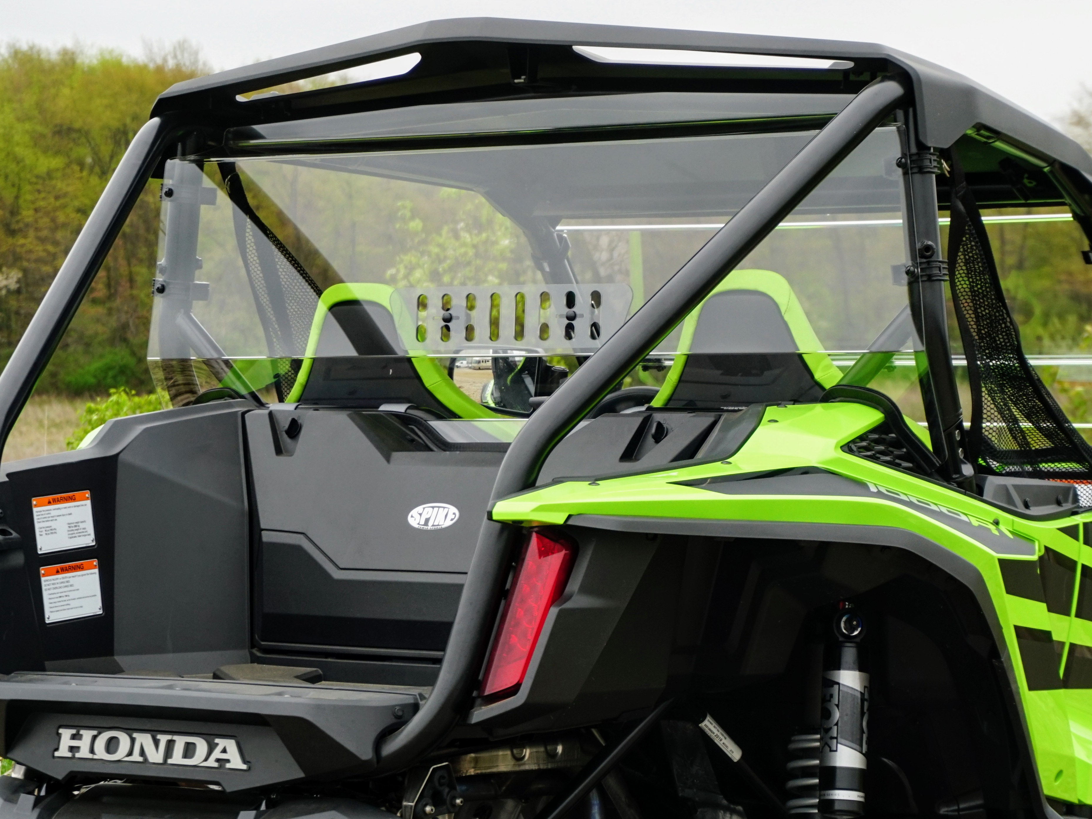 Close-up of a bright green Honda Talon with sporty bucket seats, a clear Honda Talon Rear Vented Windshield Clear-Gp 2019+, metal roll bars, and warning labels. Trees and a cloudy sky form the background.