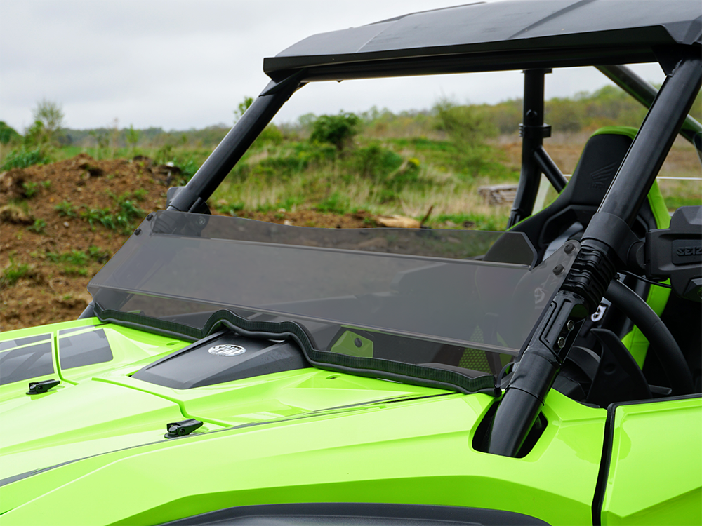 A Honda Talon 2019+ with a tinted half windshield is parked on dirt, featuring a black roof and visible seat and roll cage. The green off-road vehicle is set against grassy hills under a cloudy sky, shown from the front and side.