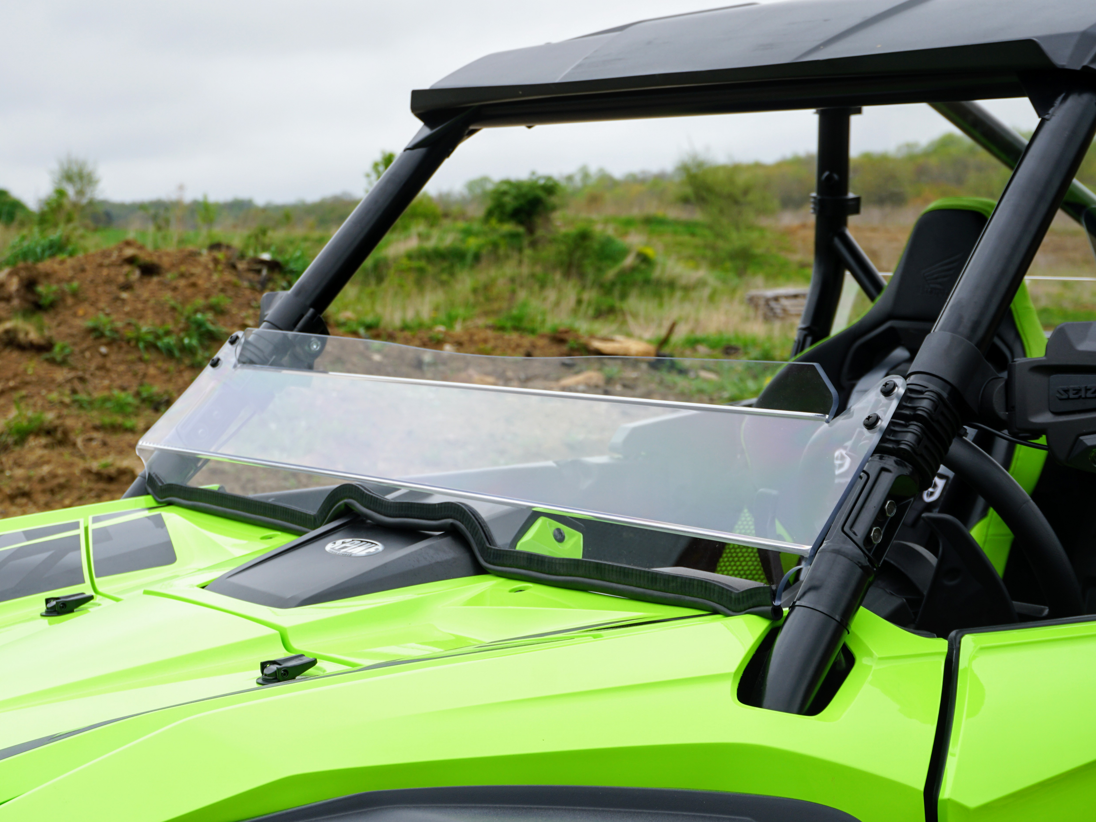 A close-up of a Honda Talon with a Hard Coated Scratch Resistant Short Shield (2019+), highlighting the windshield, front hood, and roll cage. The vehicle sits on dirt and grass with trees in the background under a cloudy sky.