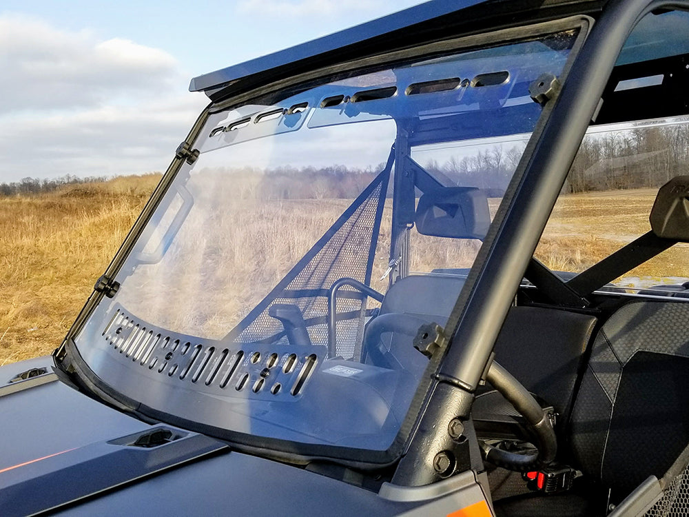 A close-up of the Polaris Ranger Full-Size 2013+ Scratch Resistant Multi-Vent Windshield by Spike, installed on a black utility vehicle in a grassy field, shows top vent slots and a visible cab interior with seats and steering wheel.