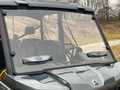 A close-up of a Can-Am Defender Comfort Flow Venting Windshield W/Hard Coat shows a clear windshield with two wipers on a gray utility vehicle cab. The vehicle is parked outside, with black seats and rural scenery in the background.