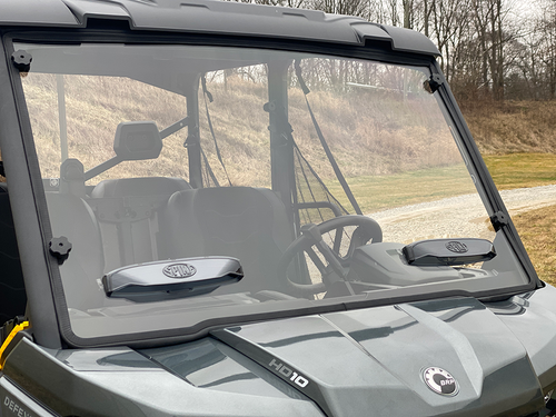 A close-up of a Can-Am Defender Comfort Flow Venting Windshield W/Hard Coat shows a clear windshield with two wipers on a gray utility vehicle cab. The vehicle is parked outside, with black seats and rural scenery in the background.