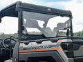 A close-up shows a 2019+ Polaris Ranger 1000/XP fitted with a Spike Powersports rear windshield, featuring the “SPIK” label. The bold “RANGER” logo is on the front, with trees and sky visible in the background.