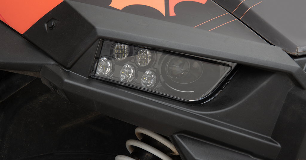 Close-up of a black vehicles angular headlight with five LED bulbs. Orange accents and fine lines decorate the body. Coiled white suspension spring is visible underneath the headlight area, indicating an off-road or ATV-style vehicle.