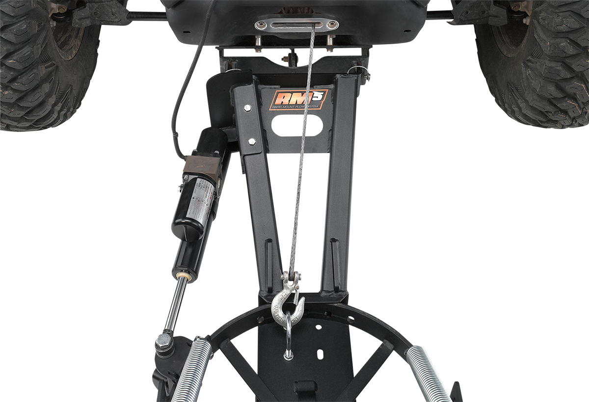 Close-up view of an ATV snow plow mount system attached under a vehicle, featuring a black metal frame, winch cable, hydraulic shock, springs, and an RM3 logo on the central bracket. The image is shot from below, showing the vehicle’s large tires.