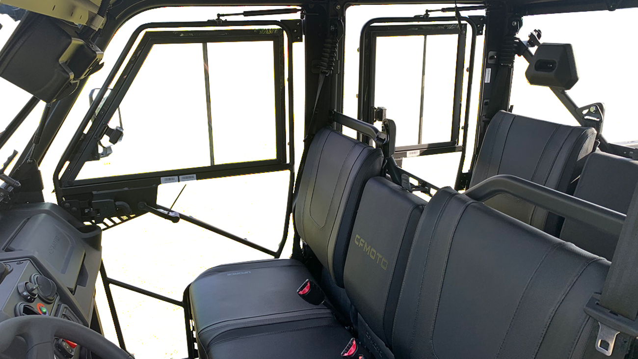 Interior view of a Can-Am off-road vehicle showing two rows of black-leather seats, seat belts, a steering wheel, and dashboard with control buttons. Large windows and doors with sturdy frames are visible, letting in ample natural light.