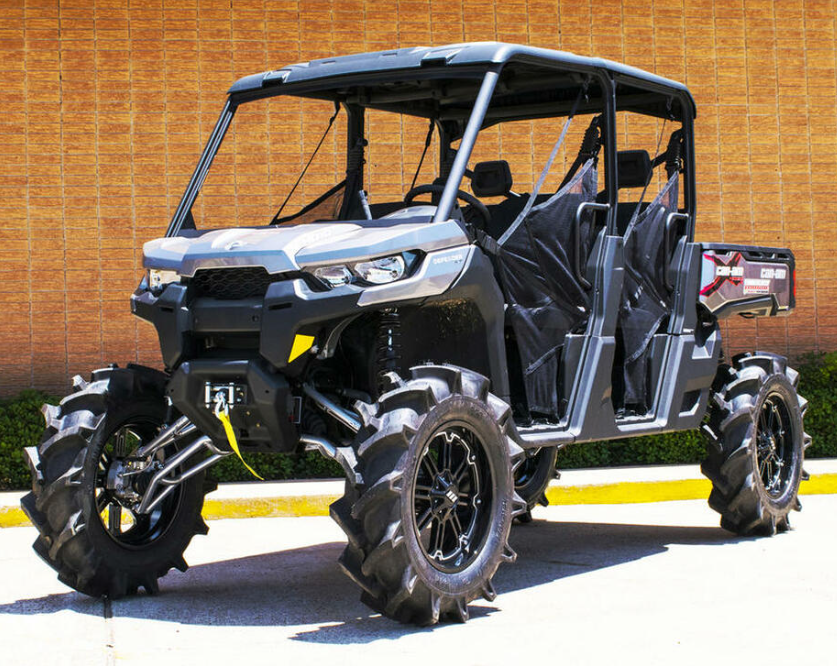 A Can-Am Defender with the S3 Power Sports 8 Lift Kit, featuring large mud tires, open mesh doors, and high ground clearance, is parked by a tan brick wall—built for rugged terrain and outdoor adventure.