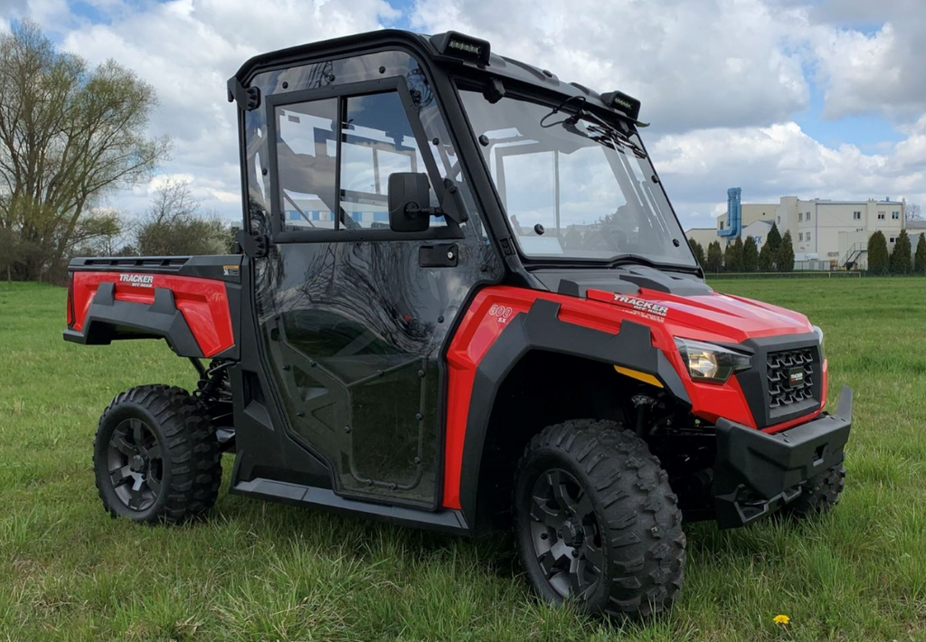 An Arctic Cat Prowler Pro/Textron Stampede 800 PREMIUM Cab Kit-equipped red and black utility vehicle with large off-road tires is parked on grass under a partly cloudy sky, featuring a glass cab with doors, mirrors, and a rear cargo bed. Trees and buildi
