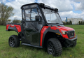 An Arctic Cat Prowler Pro/Textron Stampede 800 PREMIUM Cab Kit-equipped red and black utility vehicle with large off-road tires is parked on grass under a partly cloudy sky, featuring a glass cab with doors, mirrors, and a rear cargo bed. Trees and buildi