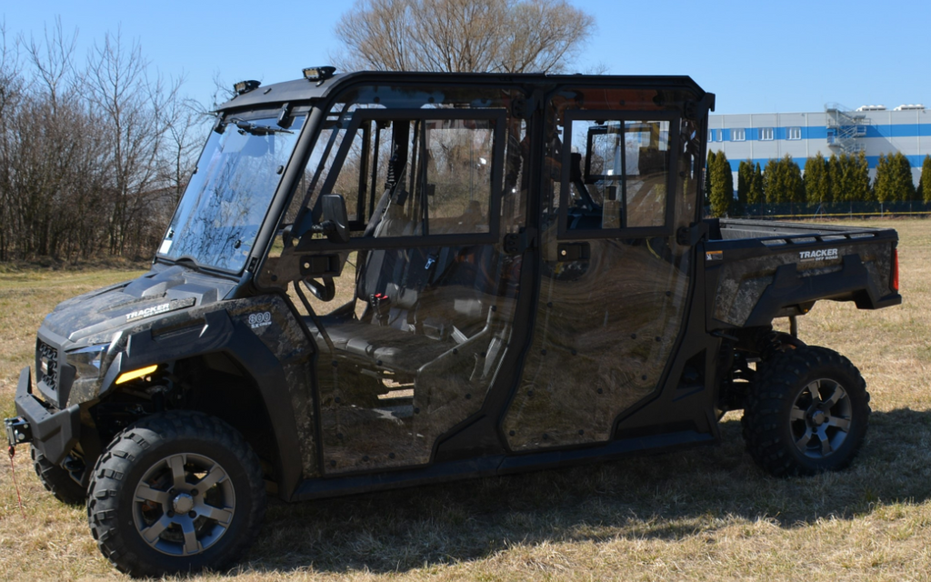 A black Arctic Cat Prowler Pro/Tracker 800 Premium Crew Cab Kit UTV with clear doors, rugged tires, and visible seating is parked on dry grass with leafless trees and a blue industrial building in the background.
