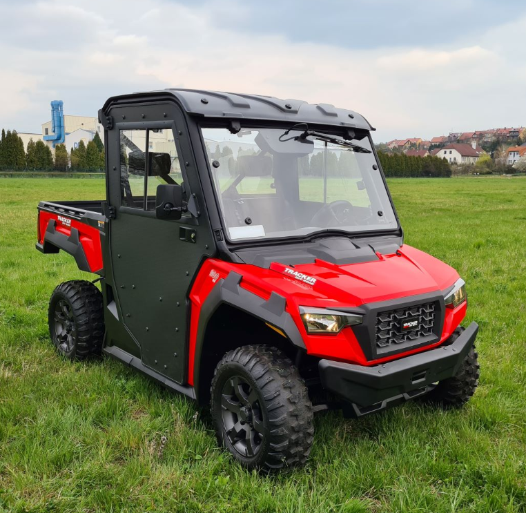 An Arctic Cat Prowler Pro 800/Textron Stampede with a Protector Cab Kit, black roof, rugged tires, side doors, sturdy front bumper, and clear windows is parked on green grass with buildings and trees in the background under a cloudy sky.