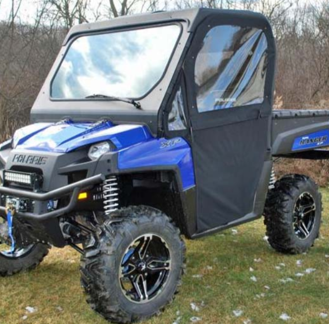 A blue Polaris Ranger 800 with Full Size Convertible Doors is parked on grass. It features large tires, a cab with doors and windows, a black roof, and rear flatbed cargo area. Trees and sparse patches of snow appear in the background.