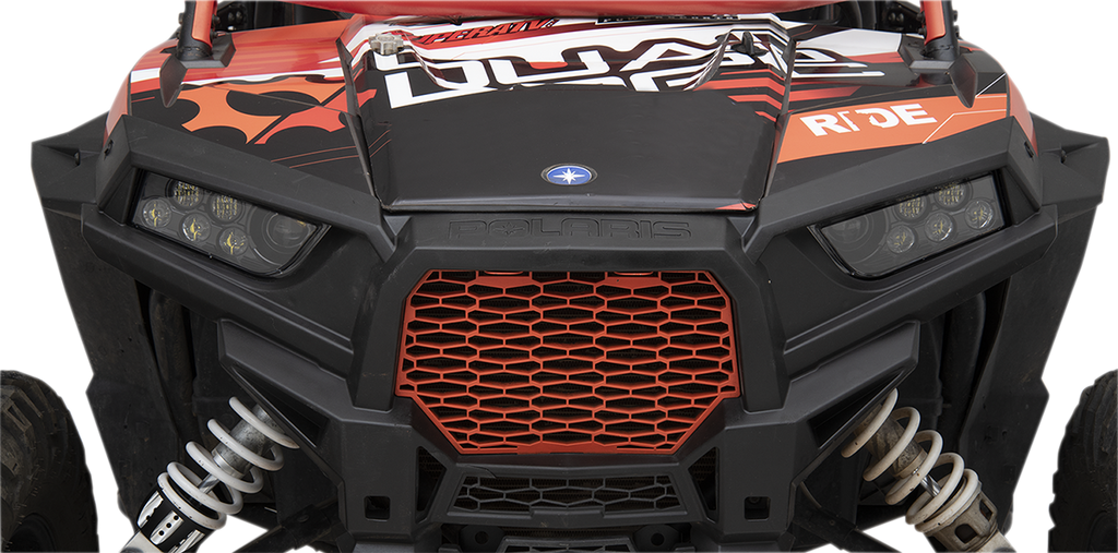 Close-up of the front of a black and red off-road vehicle with angular headlights, a honeycomb-patterned red grille, Polaris branding, coil suspension, and graphic decals including the word RIDE on the hood. The image is focused on the grille and headligh