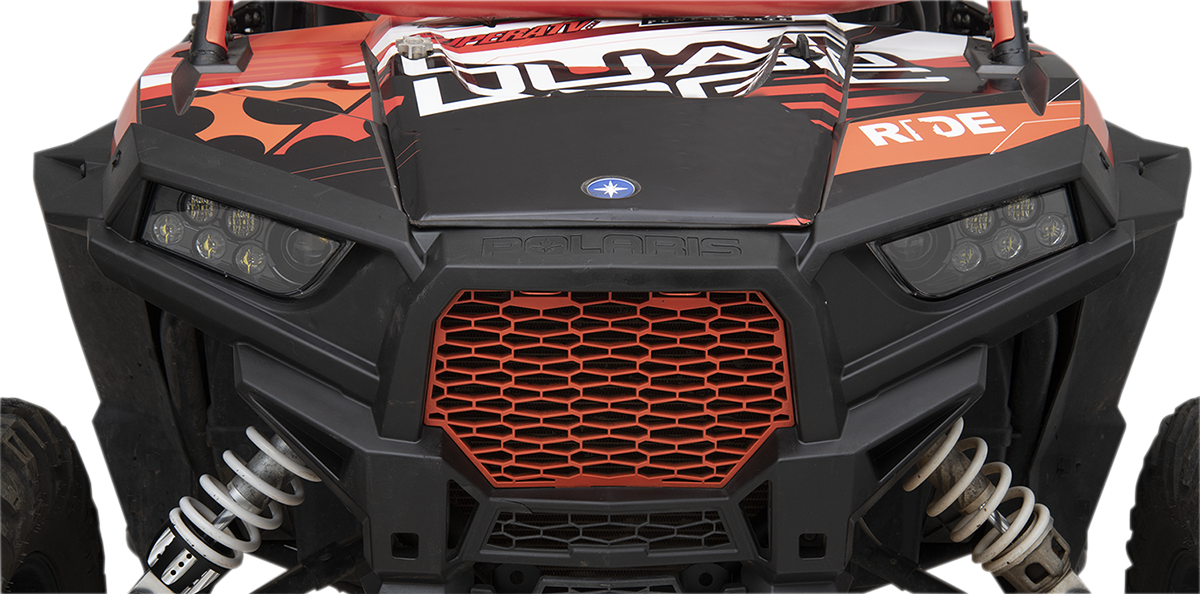 Close-up of the front of a black and red off-road vehicle with angular headlights, a honeycomb-patterned red grille, Polaris branding, coil suspension, and graphic decals including the word RIDE on the hood. The image is focused on the grille and headligh