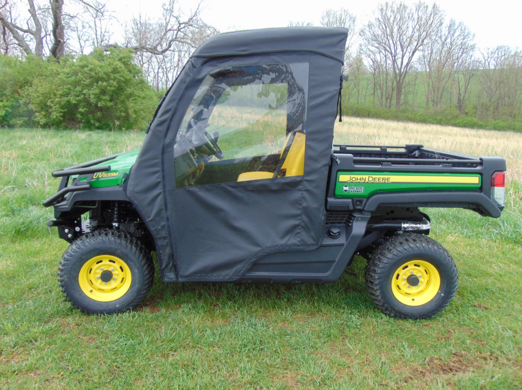 A John Deere Gator XUV with the Soft Full Door Kit is parked on grass. The zipped black soft doors and yellow wheels stand out, with trees, bushes, and an open field visible under an overcast sky in the background.
