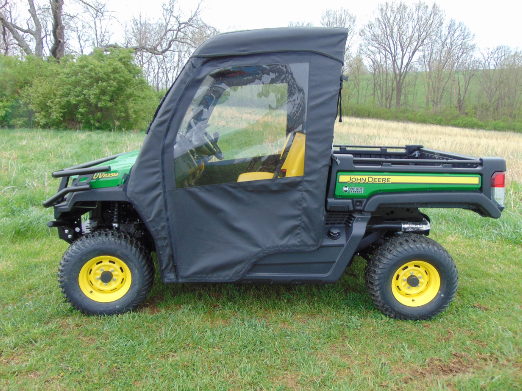 A John Deere Gator XUV with the Soft Full Door Kit is parked on grass. The zipped black soft doors and yellow wheels stand out, with trees, bushes, and an open field visible under an overcast sky in the background.