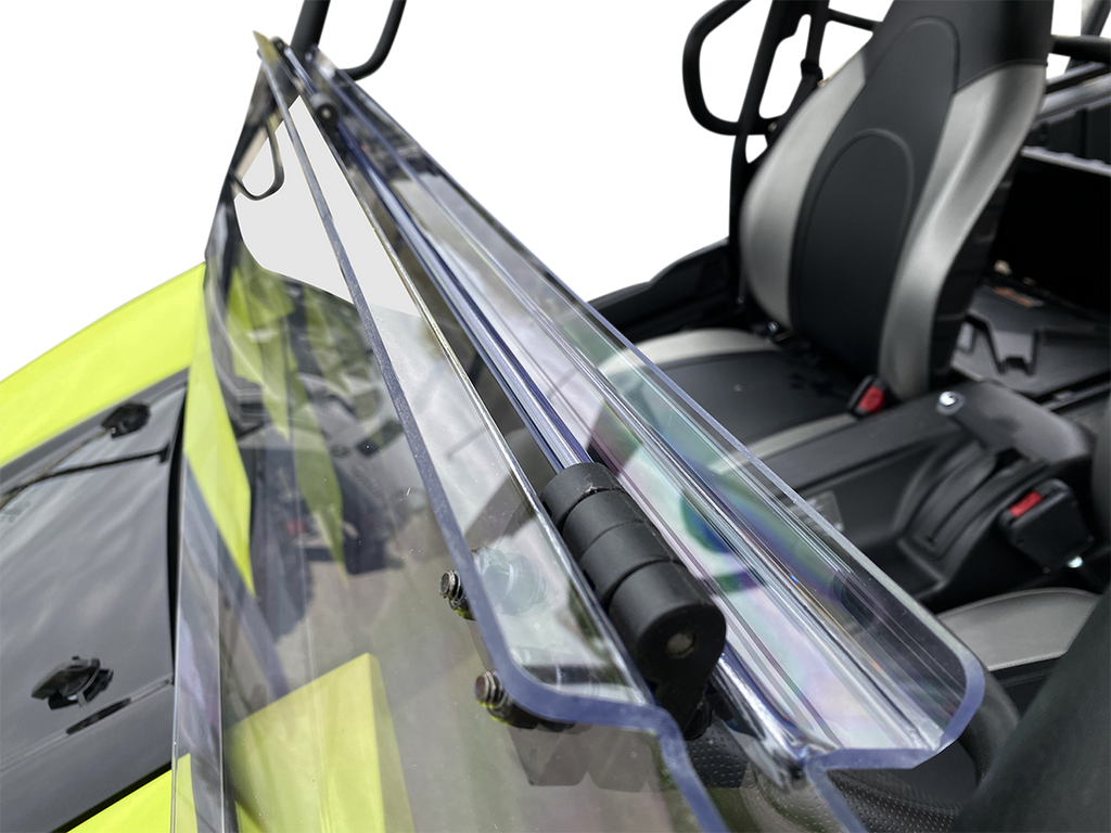 Close-up of a clear, foldable windshield attached to a black and yellow off-road vehicle. The hinge mechanism and mounting bolts are visible, and the seats and interior details of the vehicle are in the background.