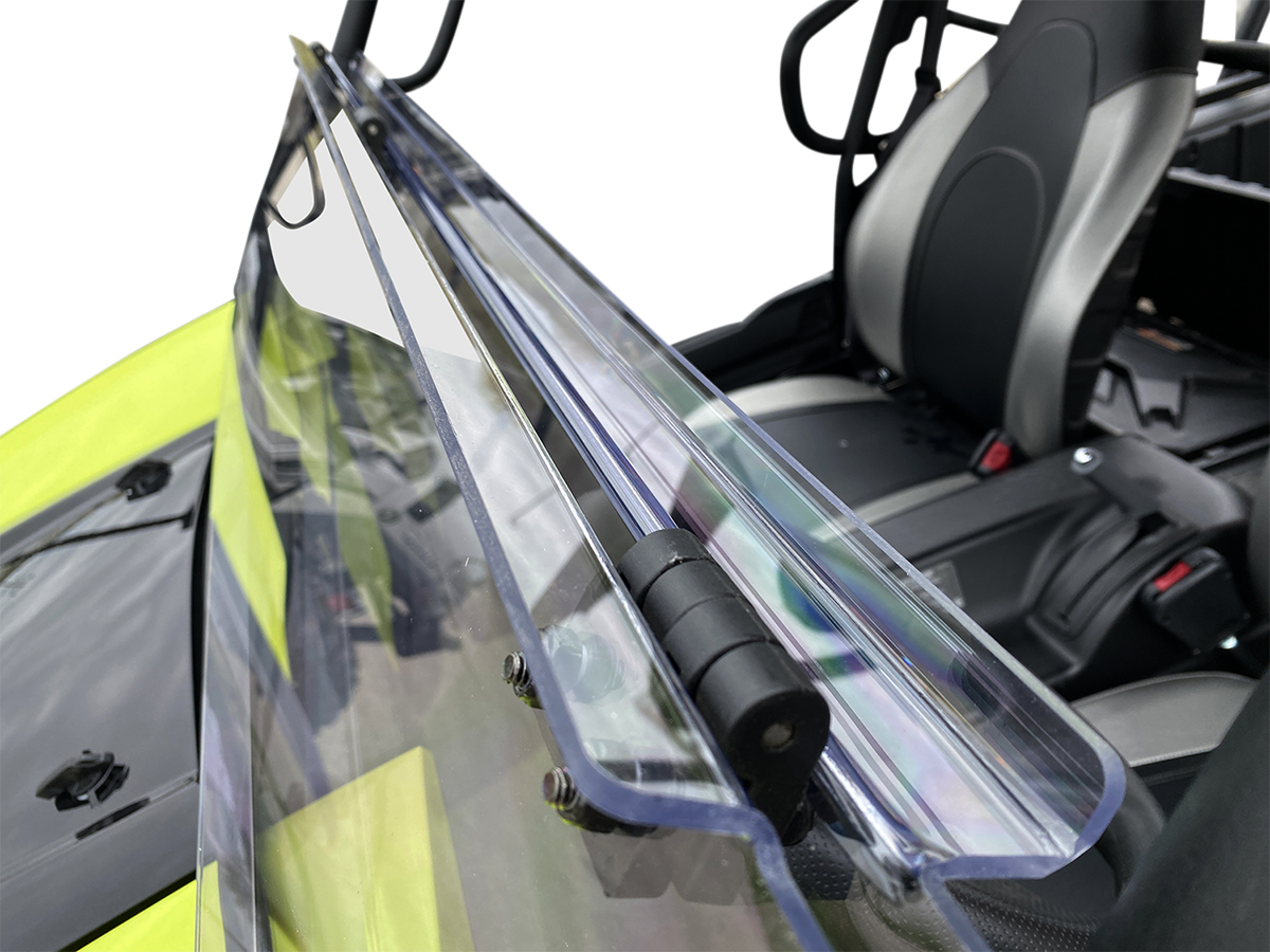 Close-up of a clear, foldable windshield attached to a black and yellow off-road vehicle. The hinge mechanism and mounting bolts are visible, and the seats and interior details of the vehicle are in the background.