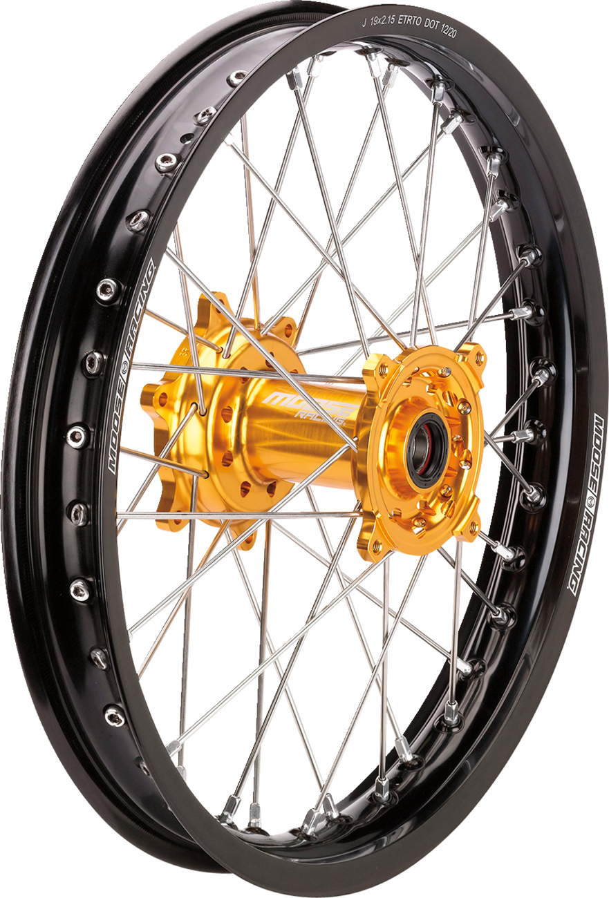 Close-up of a motorcycle wheel featuring a black rim, silver metal spokes, and a gold-colored hub. The rim displays white branding text, and the gold hub stands out with detailed machining and visible bolt holes. The background is plain white.